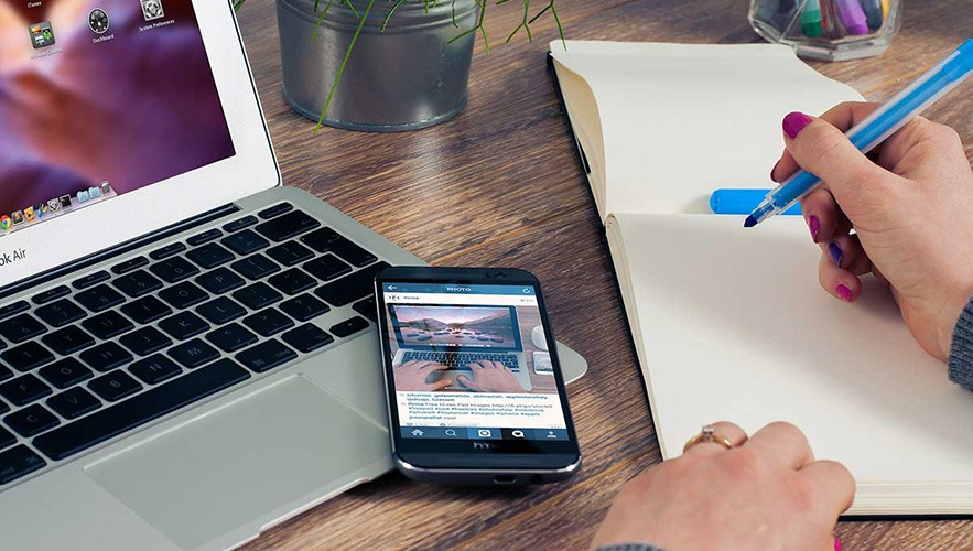 A person handles bookkeeping tasks, writing in a notebook with a blue pen beside an open laptop and smartphone on a wooden desk, with a plant and office supplies in the background.