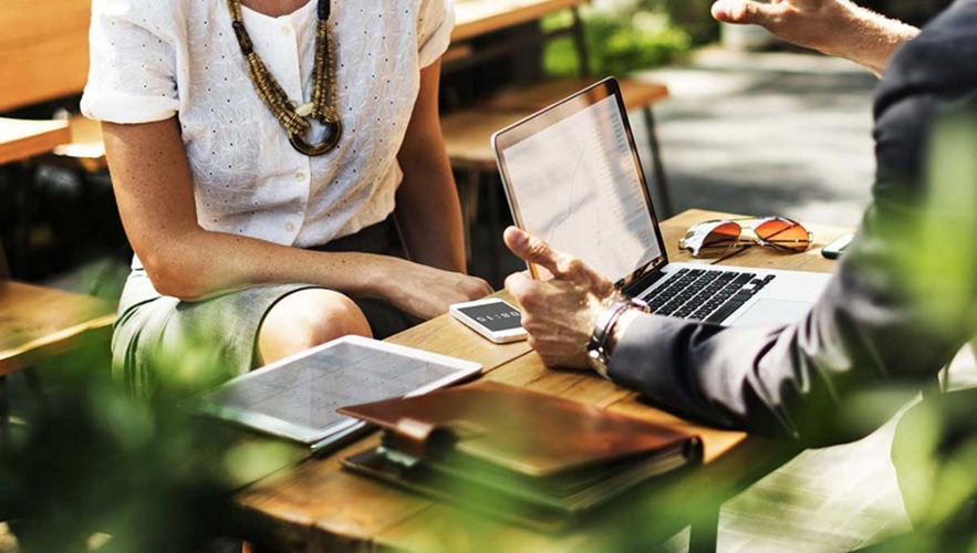 Two people sit at an outdoor table with a laptop, tablet, phone, and notebooks—discussing bookkeeping tasks. One gestures while speaking; the other listens, partially visible in a white blouse and statement necklace.