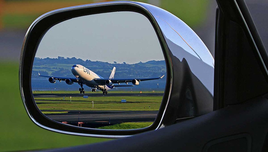 An airplane taking off is reflected in a car’s side mirror, with green fields and distant hills visible in the background—capturing a scene as precise as bookkeeping or accounting from inside the car, framed perfectly through the mirror.