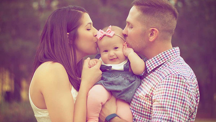 A woman and man each kiss the head of a smiling baby girl with a pink bow, who is held between them outdoors. The family appears happy and close, surrounded by soft, warm light—like a well-balanced bookkeeping or accounting record.